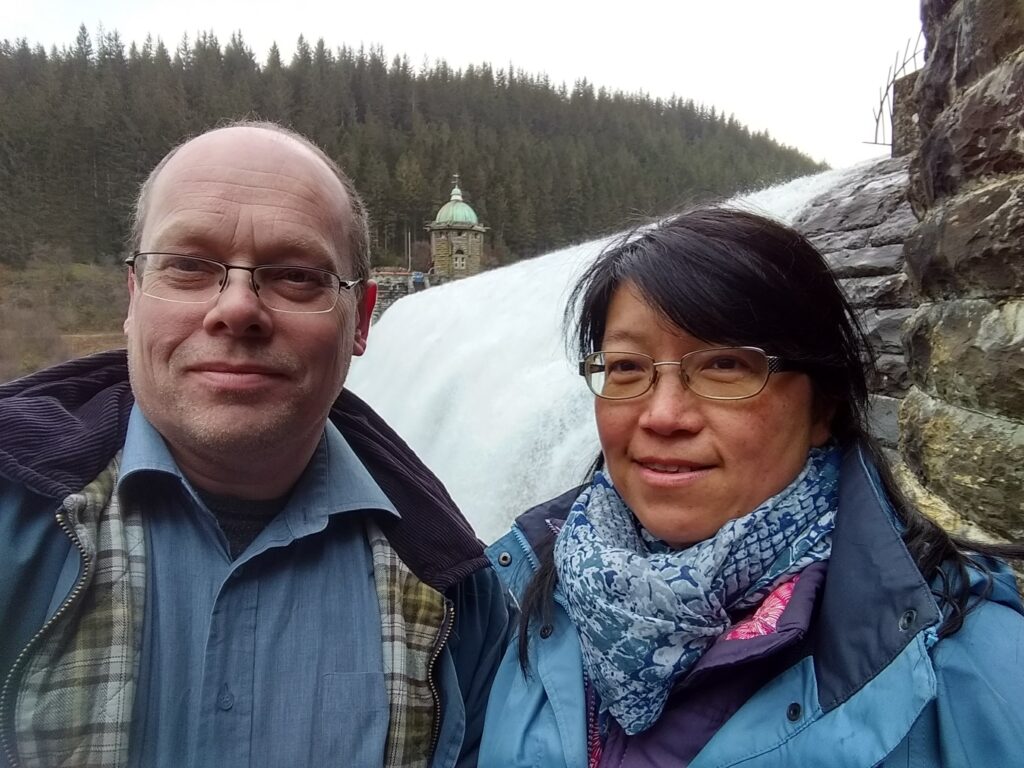 Adrian and Grace Ince stand in front of Pen y Garreg dam in the Elan Valley. Water is flowing over the top of the dam.